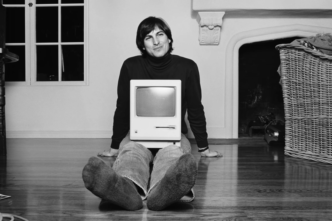 A young Steve Jobs sits on the ground with a computer on his lap.